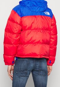 Doudoune avec un corps rouge et une capuche bleue, fabriquée en matériau brillant avec des sections quiltées horizontales. Logo de marque notable au dos.
