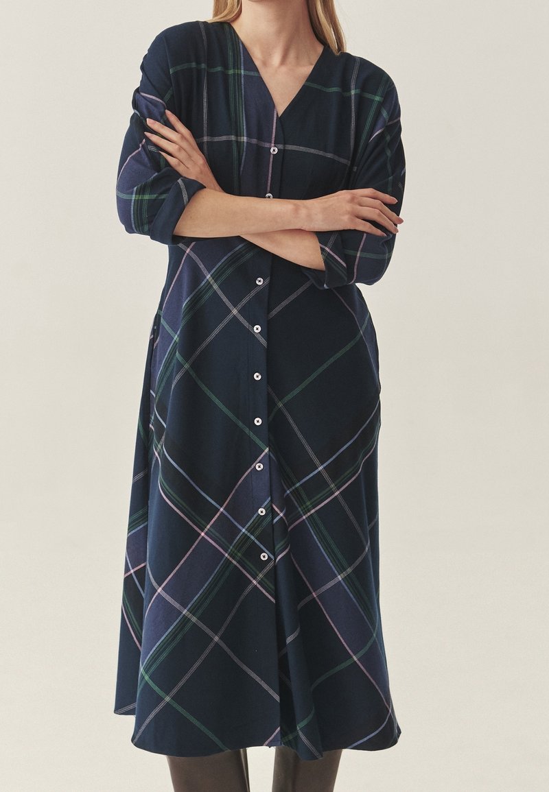 Femme portant une longue robe chemise à carreaux bleu marine avec les manches retroussées, bras croisés, debout devant un fond uni.