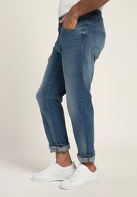 Jeans de mezclilla azul con corte tapered, puños enrollados y diseño de cinco bolsillos, combinados con zapatillas blancas. Textura suave con un ligero desgastado.