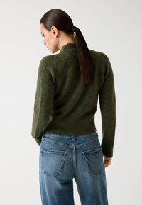 Pulover tricotat verde cu guler înalt și tiv din tricot, asortat cu blugi din denim albastru cu buzunare pe spate și etichetă logo.