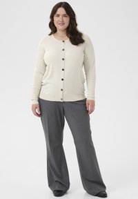 Cardigan bej deschis cu nasturi negri, manșete cu cusături, și guler rotund, asortat cu pantaloni gri largi și pantofi negri.