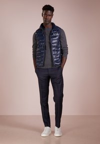 Gilet imbottito blu navy sopra un maglione grigio, abbinato a pantaloni scuri a quadri e sneakers bianche. Materiali lisci con un design aderente.