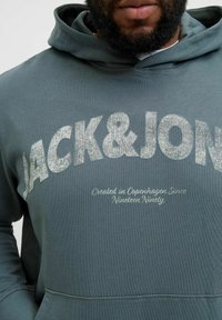 Grøn hættebluse med fedt sølvfarvet "JACK&JON" tekst og mindre hvid tekst nedenfor, lavet af et blødt, tekstureret stof.