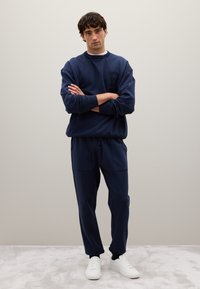 Joven con los brazos cruzados, vistiendo un sudadera azul marino y pantalones, con zapatillas blancas, de pie sobre una alfombra gris claro contra una pared lisa.