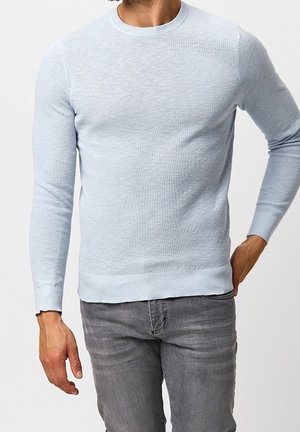Homme portant un pull à manches longues texturé bleu clair et un jean gris, debout avec une main dans la poche sur un fond uni.