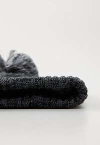 Adīta pelēka cepure ar faktūru un augšējo pom-pomu. Audumam ir mīksta un bieza izskata, ar izlocītu malu.