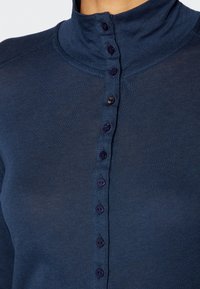 Närbild av en marinblå skjorta med hög krage, som har en vertikal rad av åtta knappar, varav en är uppknäppt nära toppen.