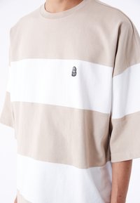 Beige ja valge triibuline lühikeste varrukatega särk, mis on valmistatud puuvillast ja mille rinnal on väike must tikandiga logo. Klassikaline ümmargune kaelus.