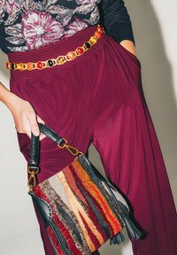 Pantaloni a gamba larga color borgogna abbinati a una borsa di stoffa con strisce multicolori e dettagli in pelle nera, chiusa da una cintura a catena dorata.