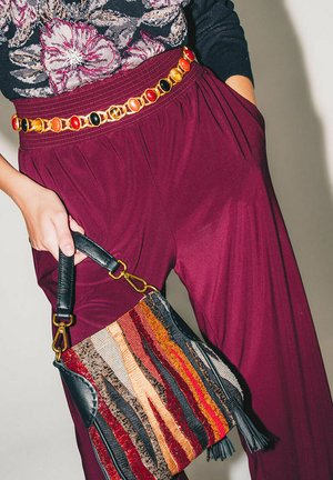 Pantaloni a gamba larga color borgogna abbinati a una borsa di stoffa con strisce multicolori e dettagli in pelle nera, chiusa da una cintura a catena dorata.