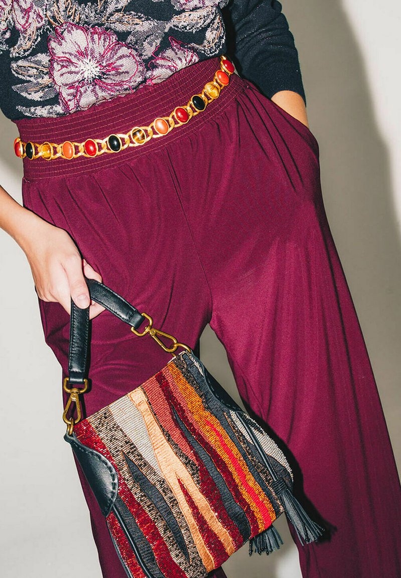 Pantaloni a gamba larga color borgogna abbinati a una borsa di stoffa con strisce multicolori e dettagli in pelle nera, chiusa da una cintura a catena dorata.