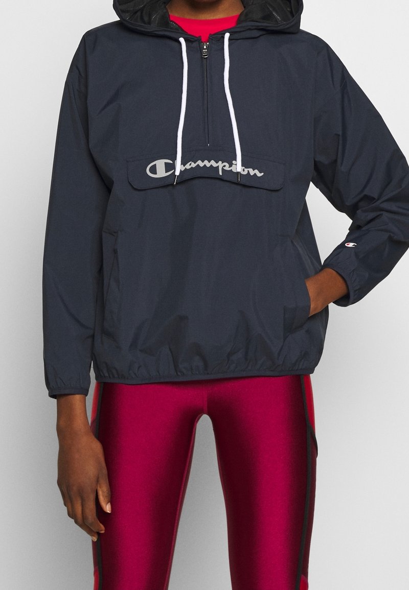 Marineblauer Windbreaker mit einer Vordertasche und einem Logo. Er verfügt über eine Kordelzugkapuze, weiße Kordeln und elastische Bündchen.