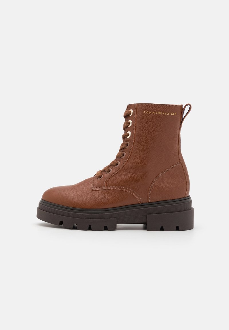 Bottine en cuir marron à lacets avec semelle épaisse noire à crampons et languette de tirage, arborant un logo "Tommy Hilfiger" doré subtil près du haut.
