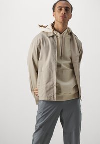 Lys beige jakke med lynlås foran, sidelommer og hætte, båret over en blød beige hættetrøje. Grå bukser fuldender outfittet.