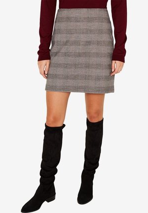 Kobieta ubrana w szarą kratkowaną mini spódniczkę, bordową bluzkę z długim rękawem i czarne zamszowe kozaki za kolano, stojąca na białym tle.