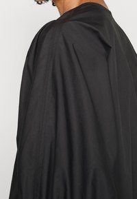 Cappotto esterno leggero e nero dal design ampio e drappeggiato con dettagli di cucitura visibili. Il tessuto appare liscio e opaco.