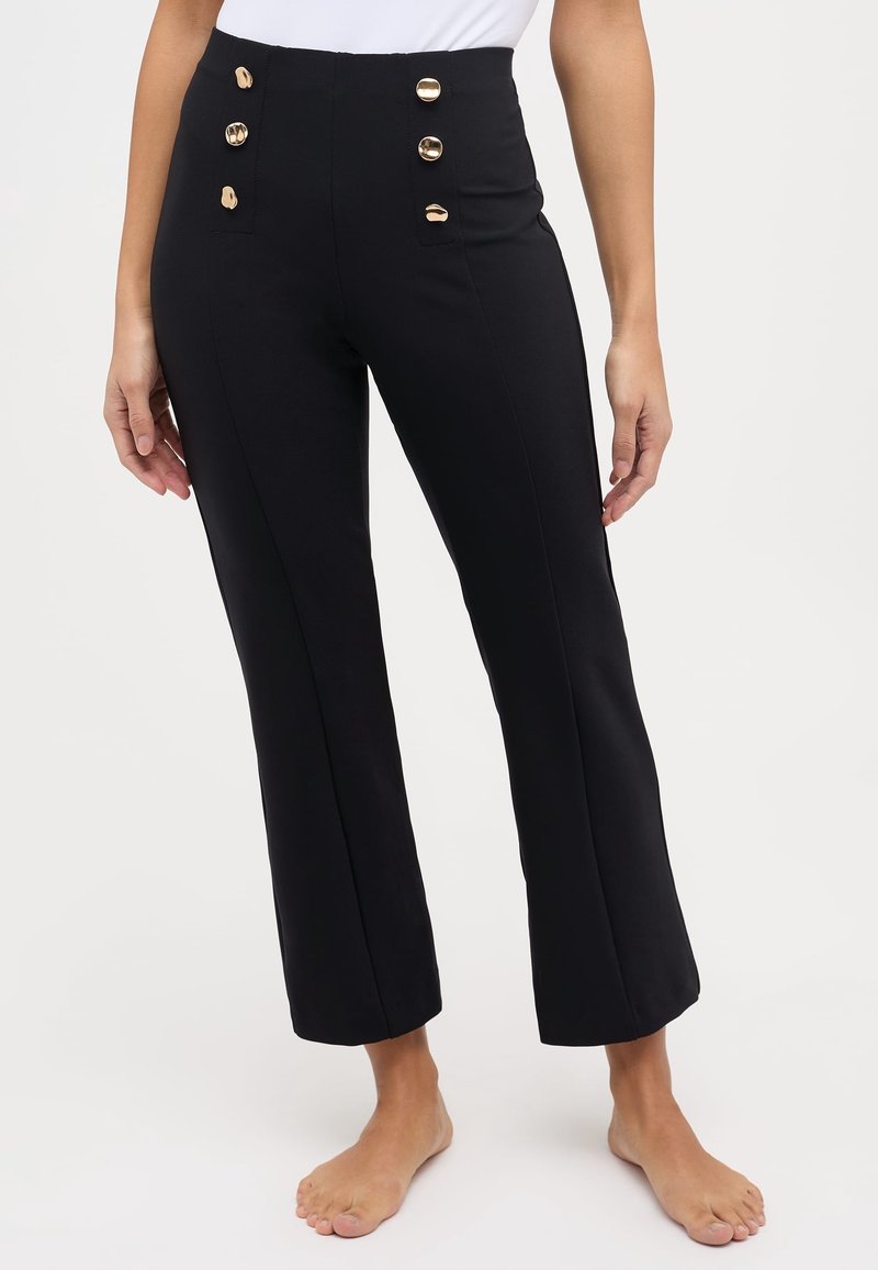Pantalon noir ajusté avec des boutons dorés à l’avant, porté par une personne pieds nus debout devant un fond uni.