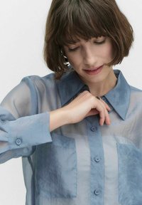 Vaaleansininen, läpikuultava nappipaita, jossa on kaksi rintataskua, pointti-kaulus ja pitkät hihat. Paidassa on hienovarainen tekstuuri ja kangasvaihtelu.