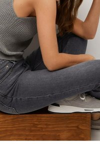 Top sin mangas de rayas grises combinado con jeans oscuros de corte ajustado y zapatillas grises claras, sentado sobre una superficie de madera.