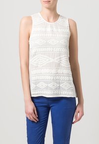 Bílý bezramínkový top s texturovanou výšivkou s geometrickými vzory. Kombinace s modrými kalhotami, které ukazují lehkou, průhlednou látku.