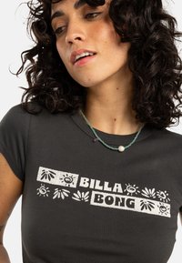 Mørkegrå t-skjorte med korte ermer, med hvit grafisk tekst "BILLA BONG" og stiliserte blomsterdesign. Enkel halslinning med perlekjede.