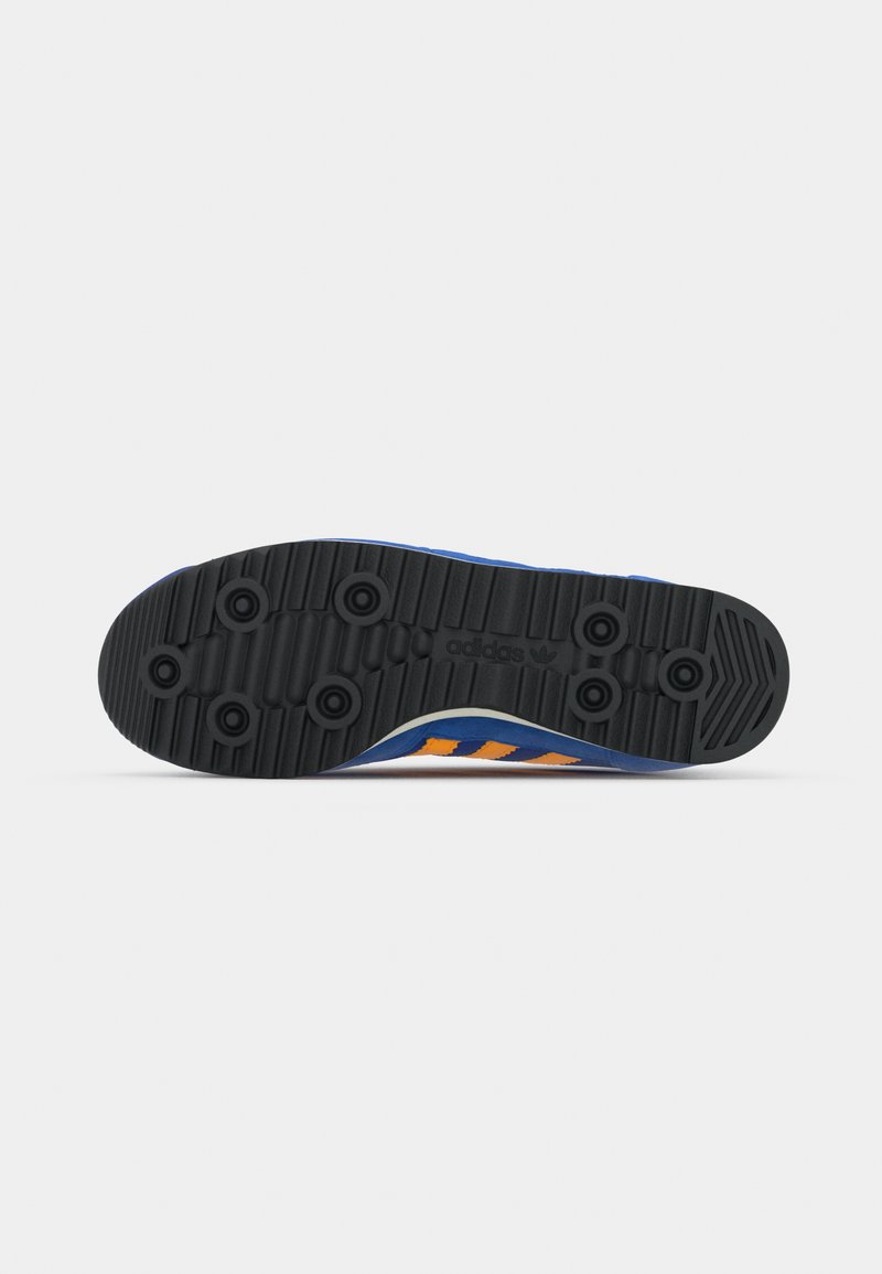 Suela de zapatilla deportiva azul con material de goma negra, que presenta patrones de banda de rodadura en forma de círculo y tres rayas naranjas. Textura suave, diseño robusto.