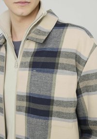 Gros plan sur une personne portant des vêtements superposés : un pull zippé beige sous une chemise à carreaux beige, marine et gris avec une poche poitrine.