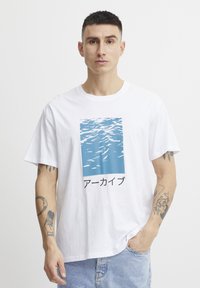 Camiseta de algodón blanca con un gráfico de ola de agua azul y texto en japonés impreso en el centro. Corte relajado con mangas cortas.