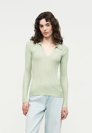 Νεαρή γυναίκα με σγουρά μαλλιά φοράει ανοιχτό πράσινο μακρυμάνικο ριμπ μπλουζάκι με γιακά και ανοιχτό μπλε τζιν σε απλό φόντο.