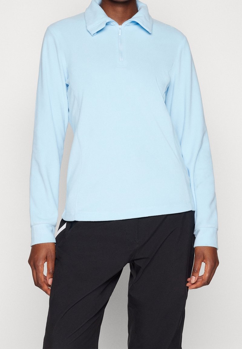 Suéter de manga larga azul claro con cuello y cremallera, hecho de tela suave. Presenta costuras laterales y un ajuste holgado. Combinado con pantalones negros.
