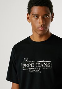 Černé bavlněné tričko s bílým texturovaným potiskem s nápisem "73 PEPE JEANS LONDON" na přední straně a kulatým výstřihem.