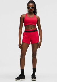 Sujetado de un sujetador deportivo rojo y pantalones cortos a juego con una cinturilla elástica negra. Zapatillas deportivas negras con suelas blancas. Persona usando gafas de sol.