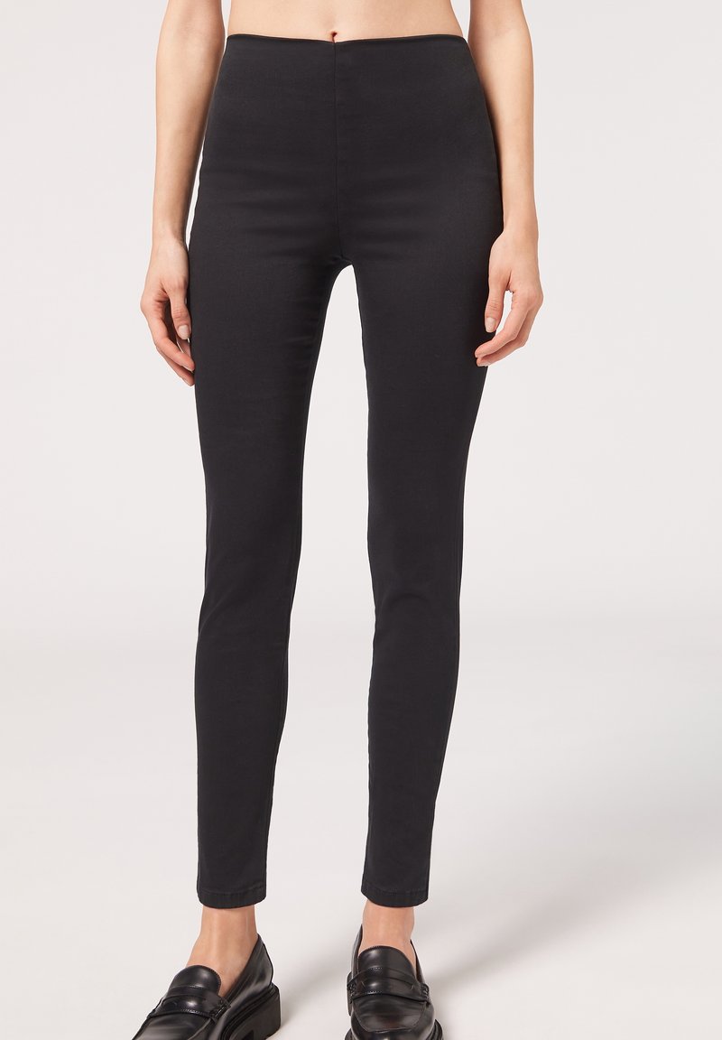 Femme portant un pantalon noir ajusté et des mocassins noirs en cuir, debout devant un fond clair uni.