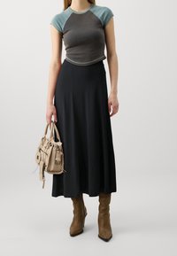 Short-sleeve, cropped top in grey/blue accent, paired with a long black skirt. Holding a beige handbag with zippers and details. Brown ankle boots.