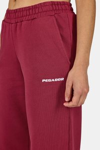 Burgundské teplákové nohavice vyrobené z mäkkej tkaniny, s elastickým pásom, bočnými vreckami a bielym logom "PEGADOR" na ľavom stehne.