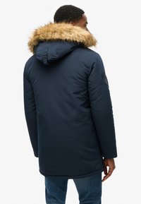 Parka blu navy con cappuccio foderato in pelliccia, design imbottito, tasche laterali e una toppa testurizzata sulla manica. Indossato sopra dei jeans in denim.