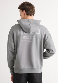 Grå hettegenser med teksturert overflate, med en stor hvit logo på ryggen og snøring i hette. Sømmer på hem og mansjetter. Løs og uformell design.