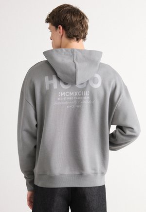 Grå hettegenser med teksturert overflate, med en stor hvit logo på ryggen og snøring i hette. Sømmer på hem og mansjetter. Løs og uformell design.