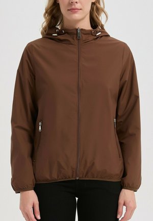 Femme portant une veste à capuche marron avec fermeture éclair, poches latérales zippées et poignets élastiques, assortie à un pantalon noir.