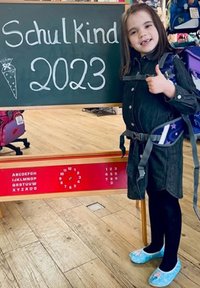 Het schoolbord toont "SchulKind 2023" met een illustratie van een wortel. Een kind in een spijkerjurk, zwarte panty’s en blauwe bloemenslippers staat ernaast.