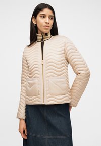 Γυναίκα που φοράει μπεζ καπιτονέ μπουφάν με φερμουάρ και τσέπες με επιδιορθώσεις, μαζί με ριγέ ζιβάγκο και σκουρόχρωμη φούστα από τζιν.