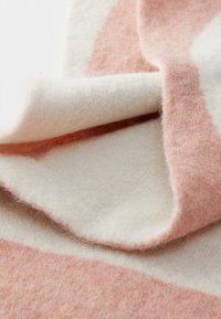 Manta têxtil com riscas suaves em creme e rosa coral, apresentando uma textura felpuda e camadas dobradas que evidenciam o seu design leve.