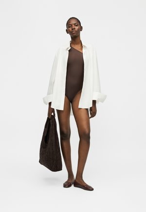 Kvinne iført brun enskuldret body, oversized hvit skjorte med knapper, brune flate sko, holder stor brun teksturert tote-veske.