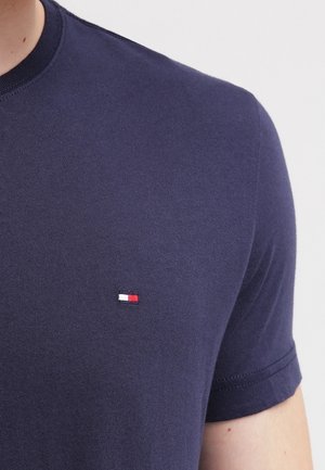 Marineblaue kurzärmelige T-Shirt mit einem kleinen rechteckigen rot-weiß-navyfarbigen Logo, das auf der linken Brust aufgestickt ist.