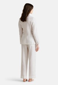 Traje de lino de color beige claro con una chaqueta ajustada y pantalones sueltos. Presenta una abertura en la espalda y un corte recto. Textura suave con un acabado limpio.