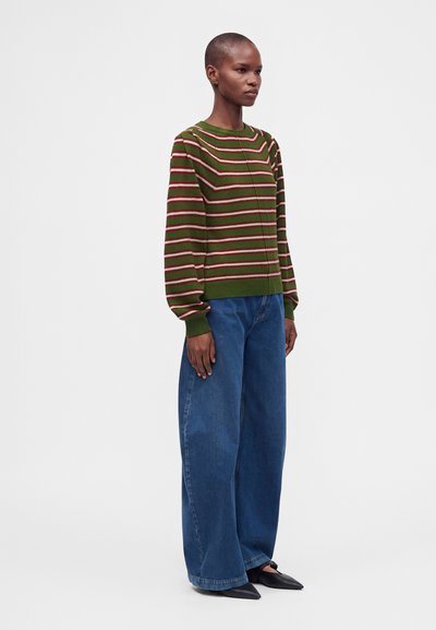 Femme aux cheveux courts portant un pull rayé vert et rouge, un jean bleu large et des chaussures noires à bout pointu, debout devant un fond uni.
