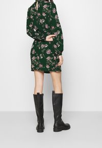 Robe fleurie verte à manches longues, taille cintrée, avec un détail dos en forme de goutte, associée à des bottes en cuir noir montantes jusqu'aux genoux.