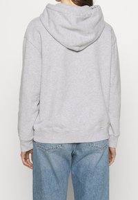 Persona che indossa un hoodie grigio chiaro e jeans blu larghi, vista da dietro su uno sfondo semplice e chiaro.