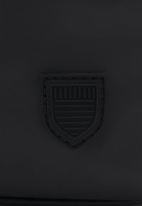 Patch noir en forme de bouclier avec des lignes embossées horizontales et verticales sur un fond de tissu foncé.