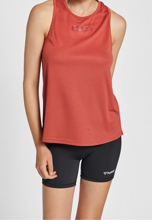 Vrouw draagt een mouwloos sporttopje in roestkleur en zwarte strakke shorts met een wit logo op de linker dij.